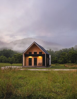 A wooden cabin with a sloped roof, a porch with a bench, and a view of a forested mountain range in the background.