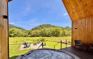 A wooden cabin with a hot tub on a deck overlooking a grassy field and hills.