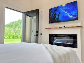 A bedroom with a bed, a TV mounted on the wall, a door leading to a balcony, and a view of greenery outside.
