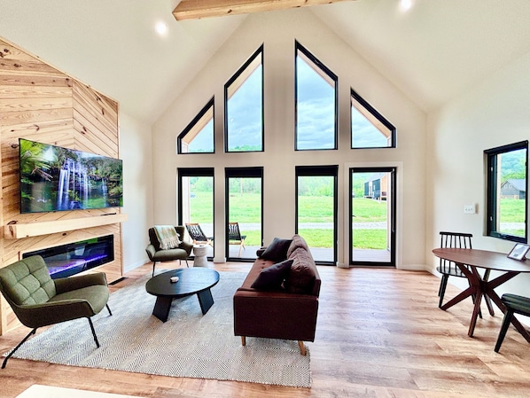 A spacious living room with a large TV, comfortable seating, a fireplace, and large windows offering views of the outdoors.