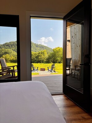 A bedroom with a large bed, a sliding glass door leading to an outdoor area with chairs and a view of a forested hill. - Knob Hill - River BEACH, Hot Tub, 360 VIEWS, Romantic (Stanton)