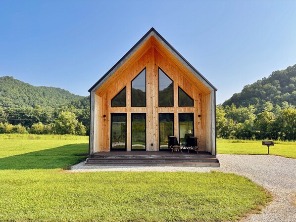 Una cabaña moderna de madera con un amplio ventanal, un porche con mobiliario y vistas a un paisaje verde con colinas al fondo.