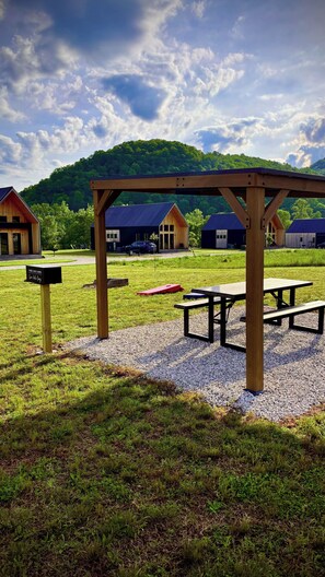 Un área de picnic con bancos y una pérgola de madera, rodeada por un campo de césped y con casas y una colina boscosa al fondo.