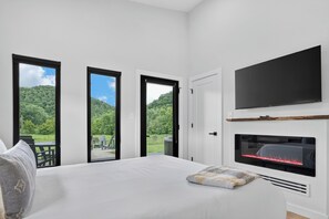 A bedroom with a bed, a flat-screen TV, a fireplace, and large windows with a view of a forested area.