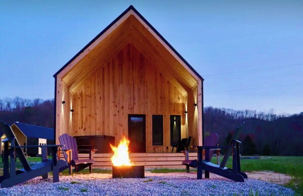 A wooden cabin with a fire pit in front, two Adirondack chairs, and a clear sky.