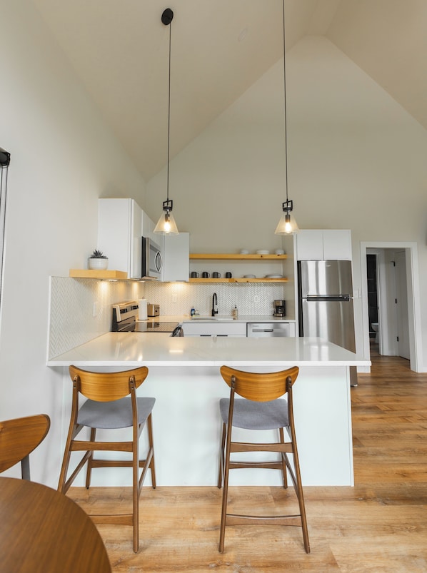 A modern kitchen with a central island, two pendant lights, wooden chairs, and a stainless steel refrigerator.