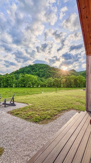 Eine Holzterrasse mit einem Kiesweg, der zu einer Bank führt, einem grünen Feld und einem bewaldeten Hügel im Hintergrund.