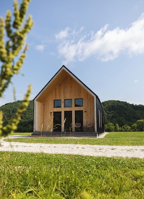 A wooden cabin with a large front door and two windows, set in a grassy field with a forested hill in the background.