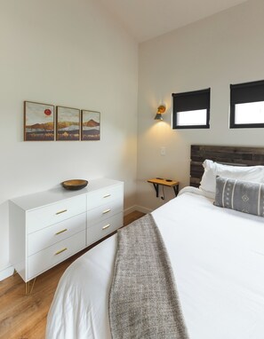 A bedroom with a bed, a dresser, a nightstand, a wall-mounted lamp, and two windows.
