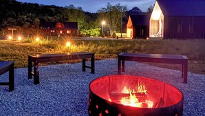 A fire pit surrounded by benches on a gravel surface, with a barn and a red building in the background.