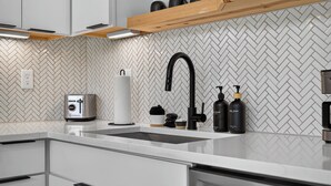 A modern kitchen with a herringbone tile backsplash, a stainless steel sink, and a wooden shelf above.