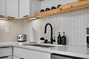 A modern kitchen with a herringbone tile backsplash, a stainless steel sink, and a wooden shelf above.