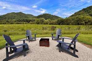 A seating area with four Adirondack chairs arranged around a fire pit on a gravel surface, set against a backdrop of lush green hills and a clear sky.