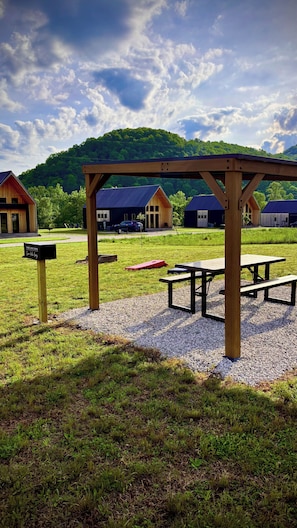 A picnic area with benches and a wooden pergola, surrounded by a grassy field and a backdrop of houses and a forested hill.