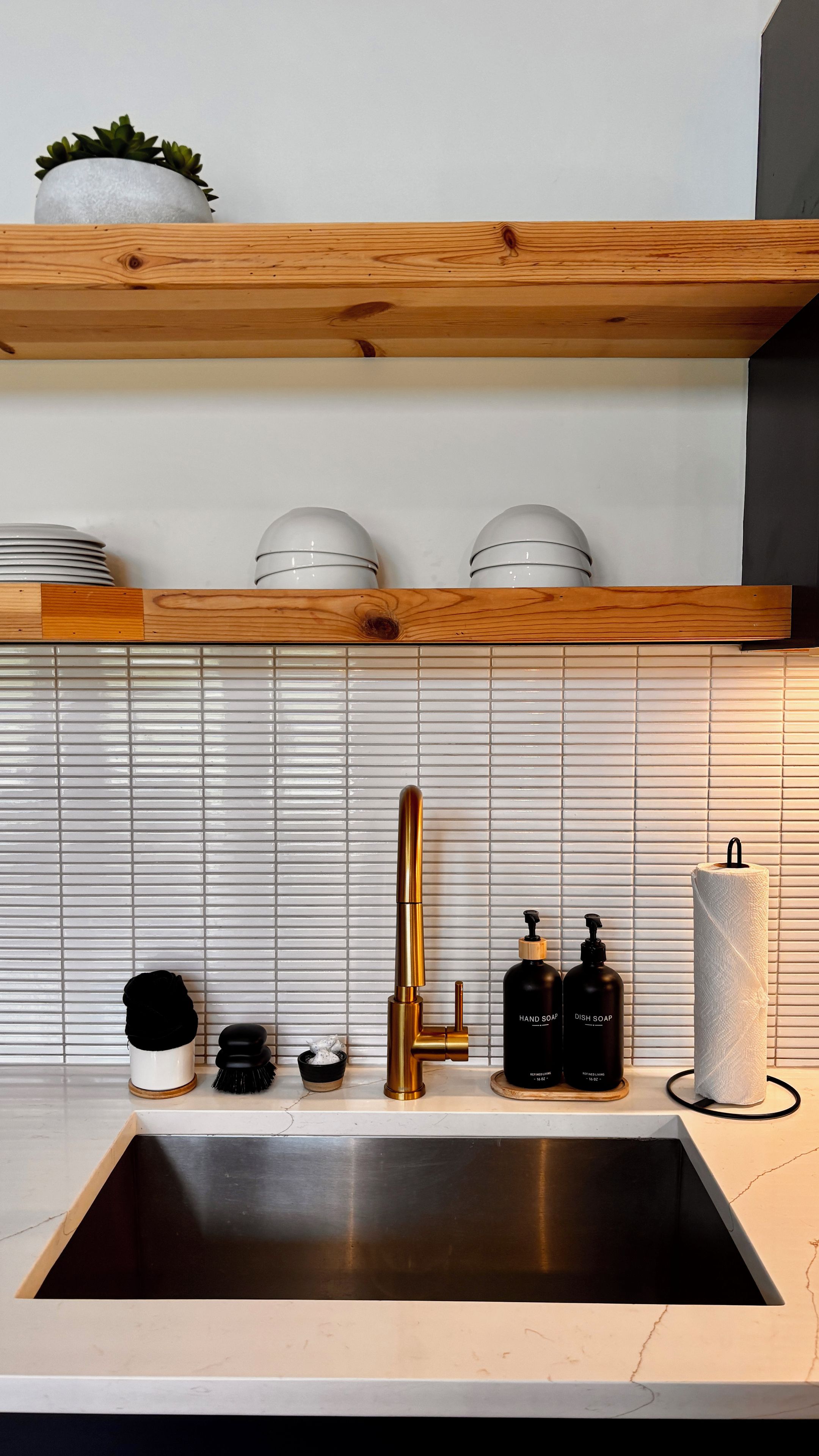 A modern kitchen with a stainless steel sink, a gold faucet, and a wooden countertop. There are two white spherical objects on a shelf above the sink.
