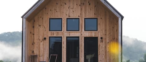 Una cabaña de madera con techo a dos aguas, tres ventanas y un porche con dos sillas y una mesa.
