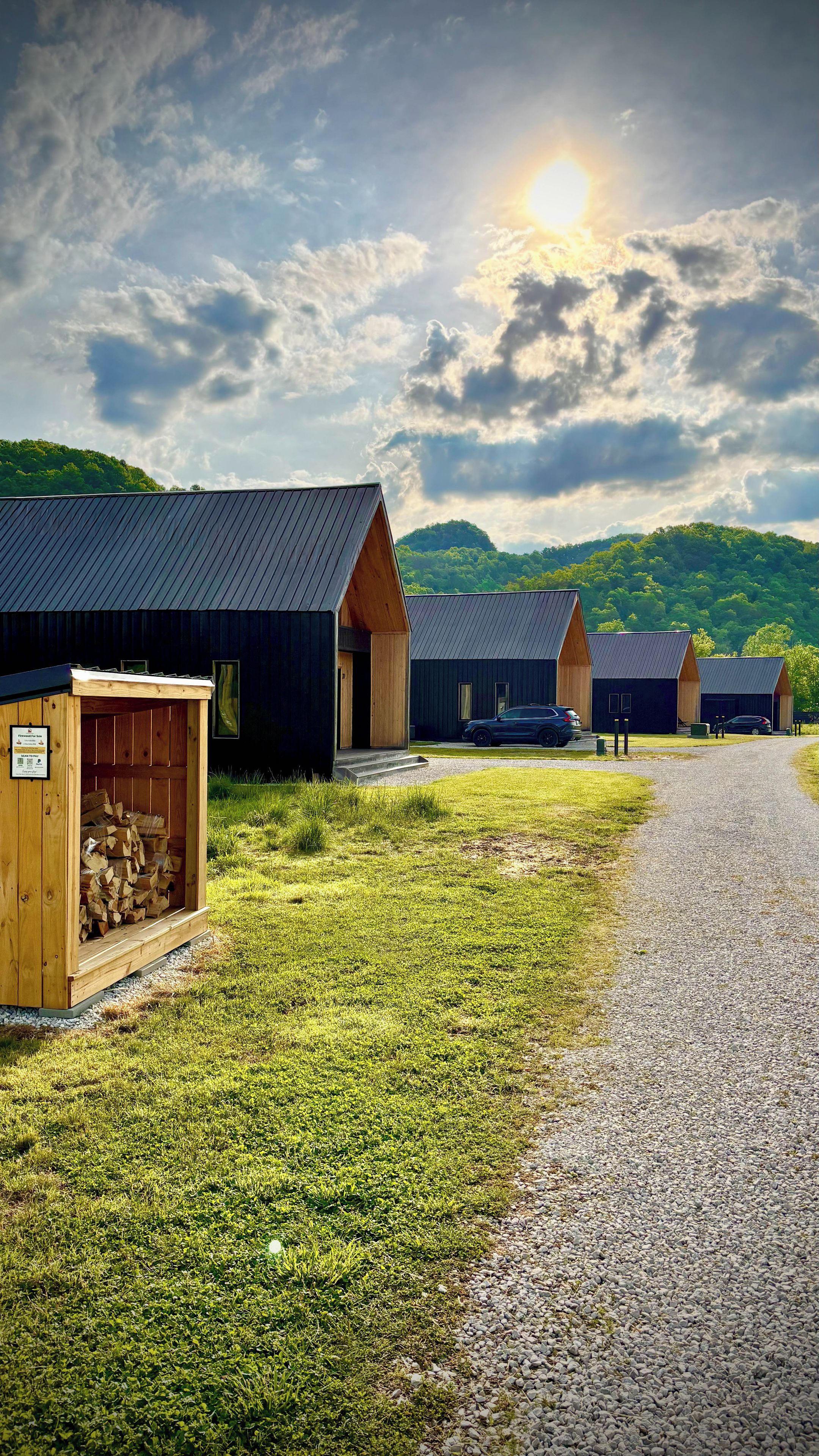 A row of modern cabins with a gravel path leading to them, set against a backdrop of green hills and a cloudy sky.