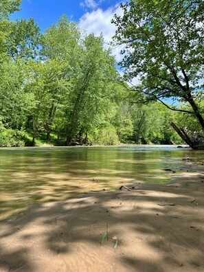 A riverbank with clear water, surrounded by dense green foliage and trees. - Perfect Perch - RARE River with BEACH, Sunsets and Stars, Hot Tub, Mountain View (Stanton)