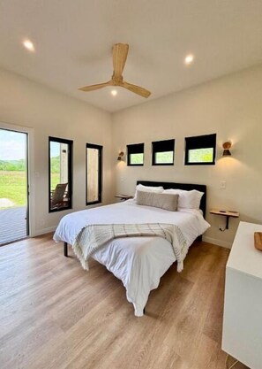 A bedroom with a large bed, a ceiling fan, and a view of a green landscape through a sliding door.