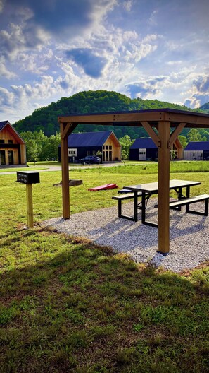 Un espace pique-nique équipé de bancs et d’une pergola en bois, entouré d’un champ verdoyant et avec, en arrière-plan, des maisons et une colline boisée.