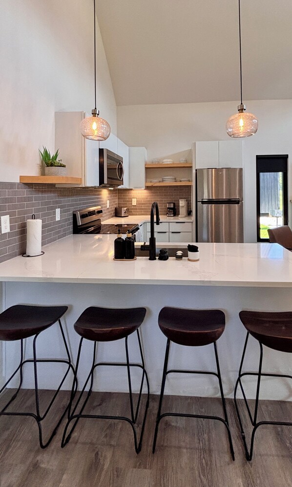 Une cuisine moderne dotée d’un îlot central, de tabourets de bar, d’appareils électroménagers en acier inoxydable et de luminaires suspendus.