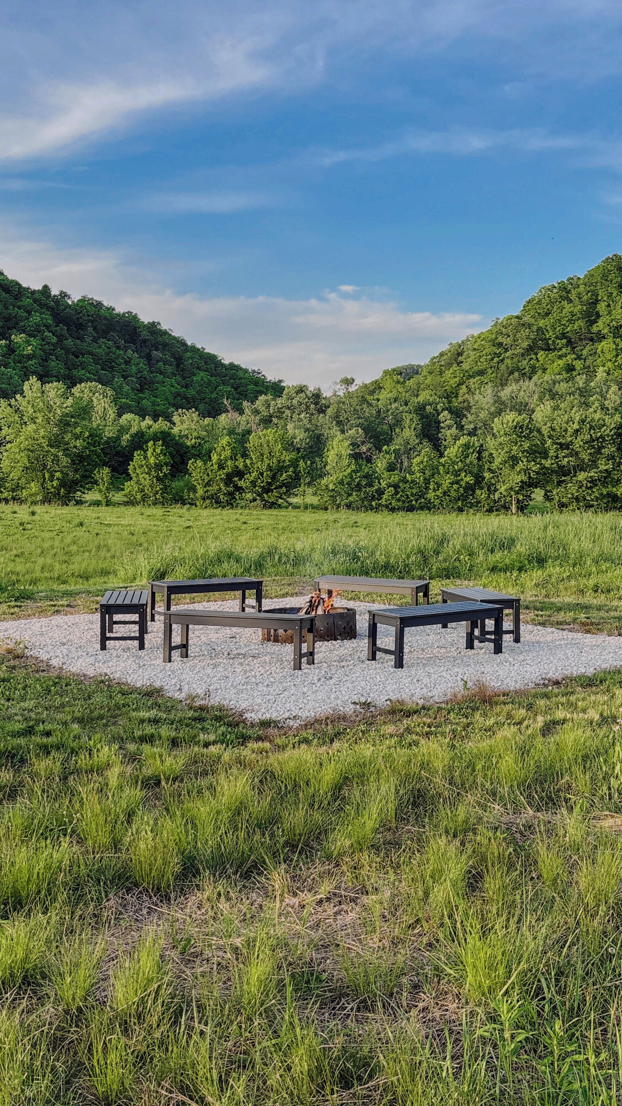 A campsite with four wooden benches arranged in a circle around a central fire pit, set in a grassy field with a forested hill in the background.