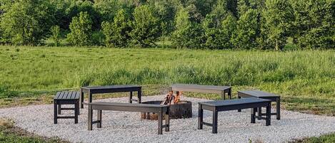 A campsite with four wooden benches arranged in a circle around a central fire pit, set in a grassy field with a forested hill in the background.