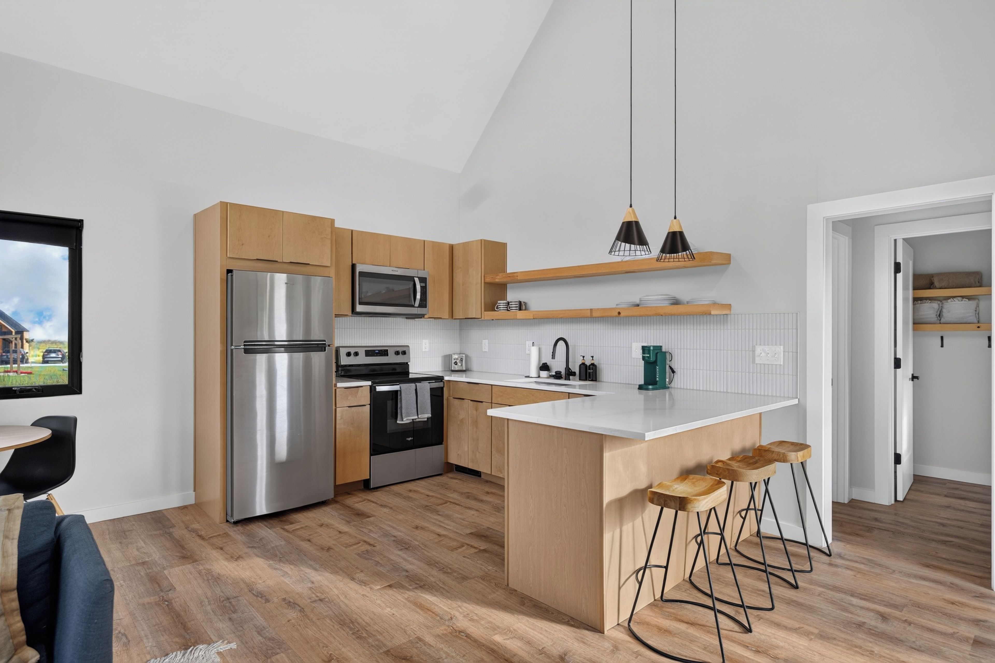 A modern kitchen with a stainless steel refrigerator, a microwave, and an oven. There are wooden cabinets, a white countertop, and a bar stool area.