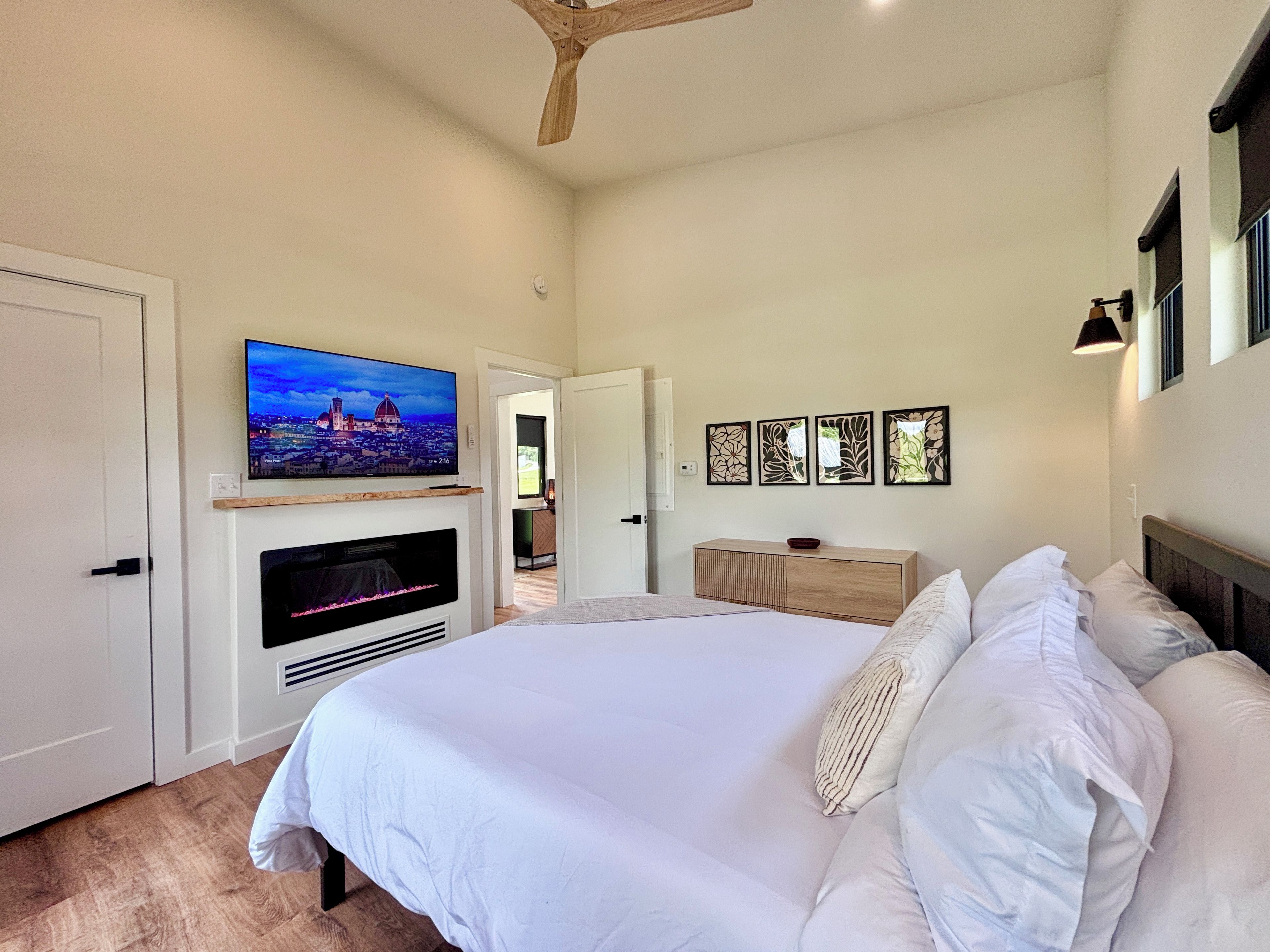 A bedroom with a bed, a television mounted on the wall, a fireplace, and a door leading to another room.