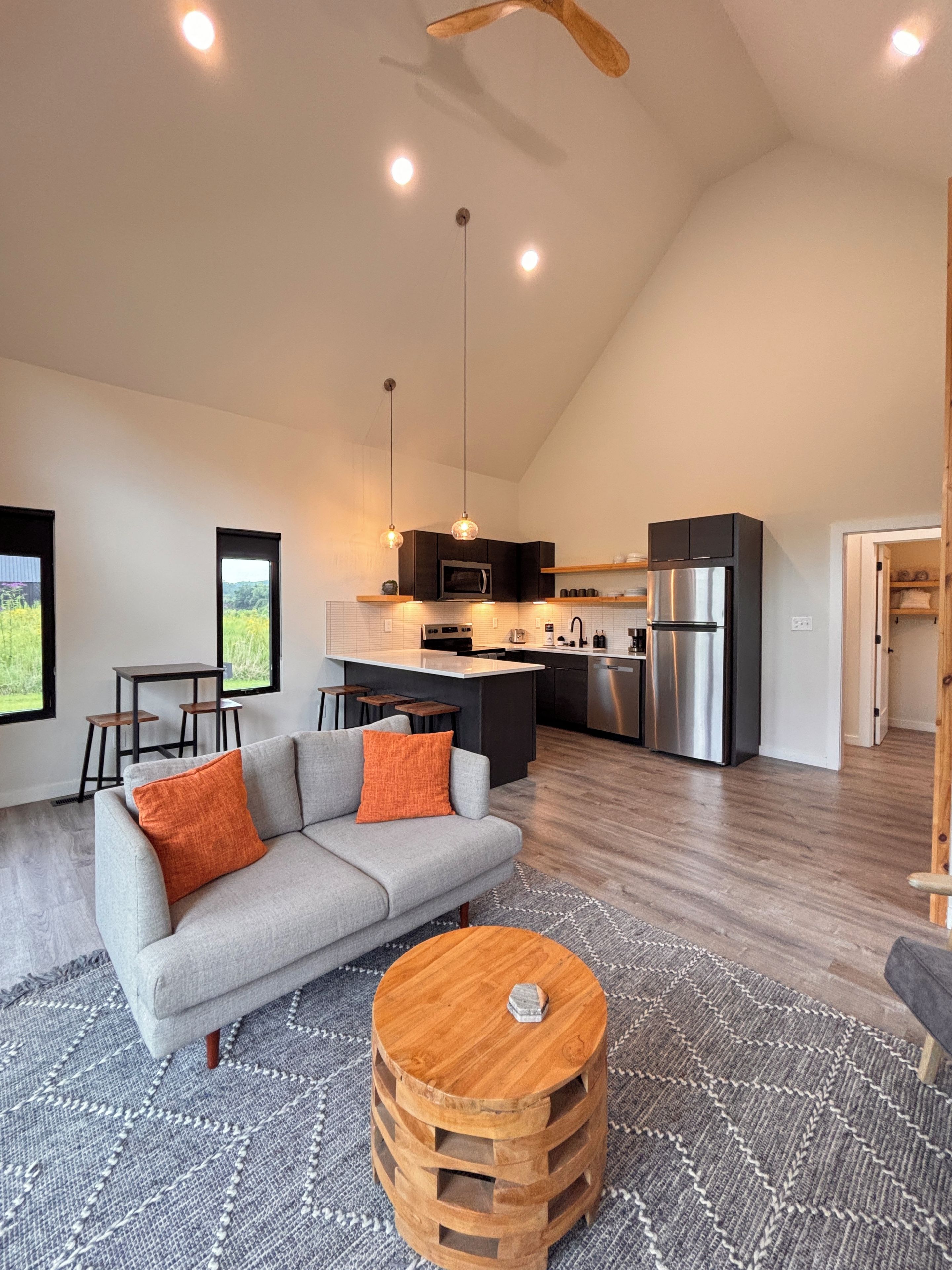 A modern kitchen with a central island, a dining area with a gray sofa and wooden stools, and a large window offering a view of greenery.