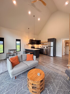 A modern kitchen with a central island, a dining area with a gray sofa and wooden stools, and a large window offering a view of greenery. - Moonrise Meadow | RARE River BEACH, Hot Tub, RRG (Stanton)