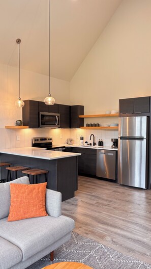 Une cuisine moderne dotée d’un îlot central, de meubles de couleur sombre, d’appareils électroménagers en acier inoxydable et de luminaires suspendus.