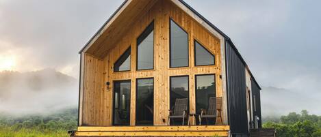 A wooden cabin with large windows, a porch, and a view of a grassy field.