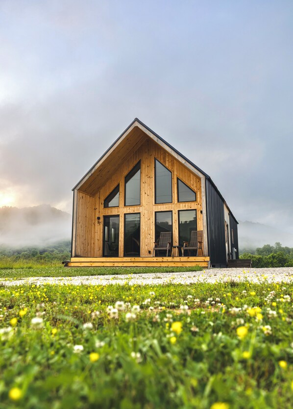 A wooden cabin with large windows, a porch, and a view of a grassy field.