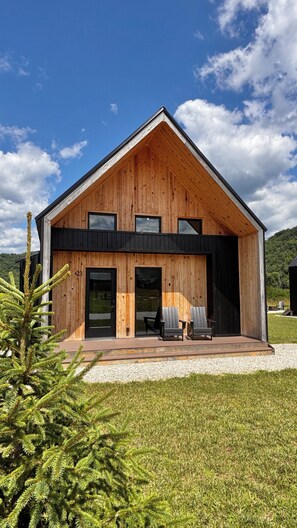 A modern wooden house with a sloped roof, large windows, and a covered porch with outdoor seating.