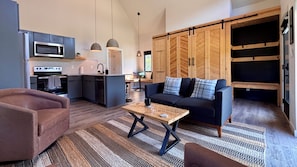 A modern kitchen with a microwave, stove, and dishwasher. There is a dining area with a table and chairs. The room has a large wooden wardrobe and a sofa with cushions. - River Rascal - Scenic views, River with BEACH, Hot Tub, Fire pit, Starry Nights (Stanton)