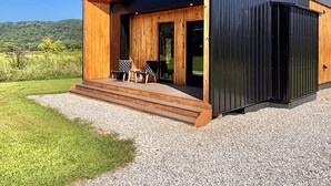 A modern wooden house with a large porch, two visible windows, and a gravel driveway.