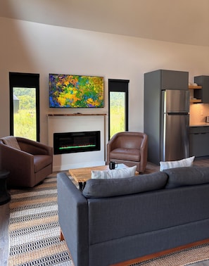A modern living room with a sofa, armchairs, a flat-screen TV, a fireplace, and a kitchen area. - River Rascal - Scenic views, River with BEACH, Hot Tub, Fire pit, Starry Nights (Stanton)