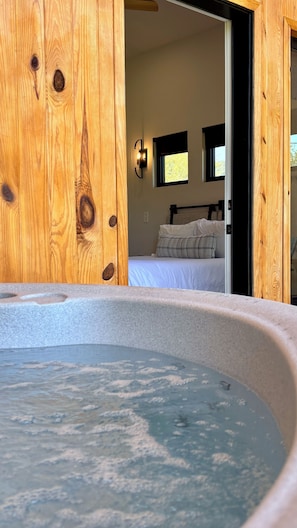 A room with a wooden door, a bed with white bedding, a nightstand, and a wall-mounted light fixture.
