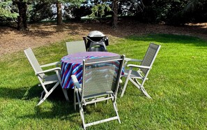 A table with a colorful tablecloth, surrounded by four metal chairs, set on a grassy lawn with trees in the background.