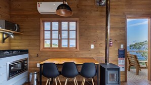 A cabin interior with a kitchen, dining area, and a large window offering a view. - Cabana c\ lareira prox. de Florianópolis VST033 (Anitapolis)