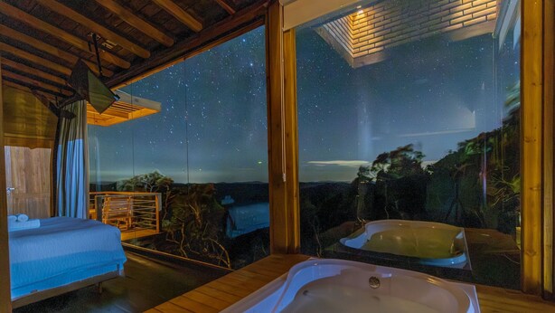 A room with a large window offering a starry night view, a wooden ceiling, and a bathtub. - Cabana c\ lareira prox. de Florianópolis VST033 (Anitapolis)