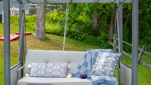 A swing set with a cushioned seat and two pillows, set in a grassy outdoor area with a wooden fence and greenery in the background.