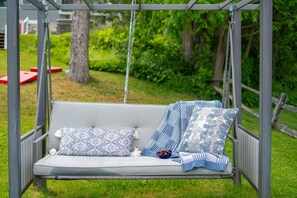 A swing set with a cushioned seat and two pillows, set in a grassy outdoor area with a wooden fence and greenery in the background. - Hot Tub, 5 min to Colgate, Sleeps 6, Welcome Wine (Madison)