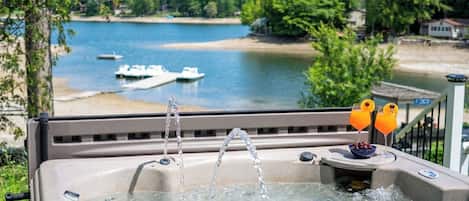 A hot tub with two faucets, a small table with two drinks, and a view of a lake with boats and a beach.