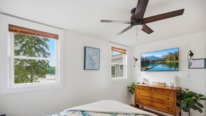 A bedroom with a bed, a dresser, a ceiling fan, and a large window with a view of trees.