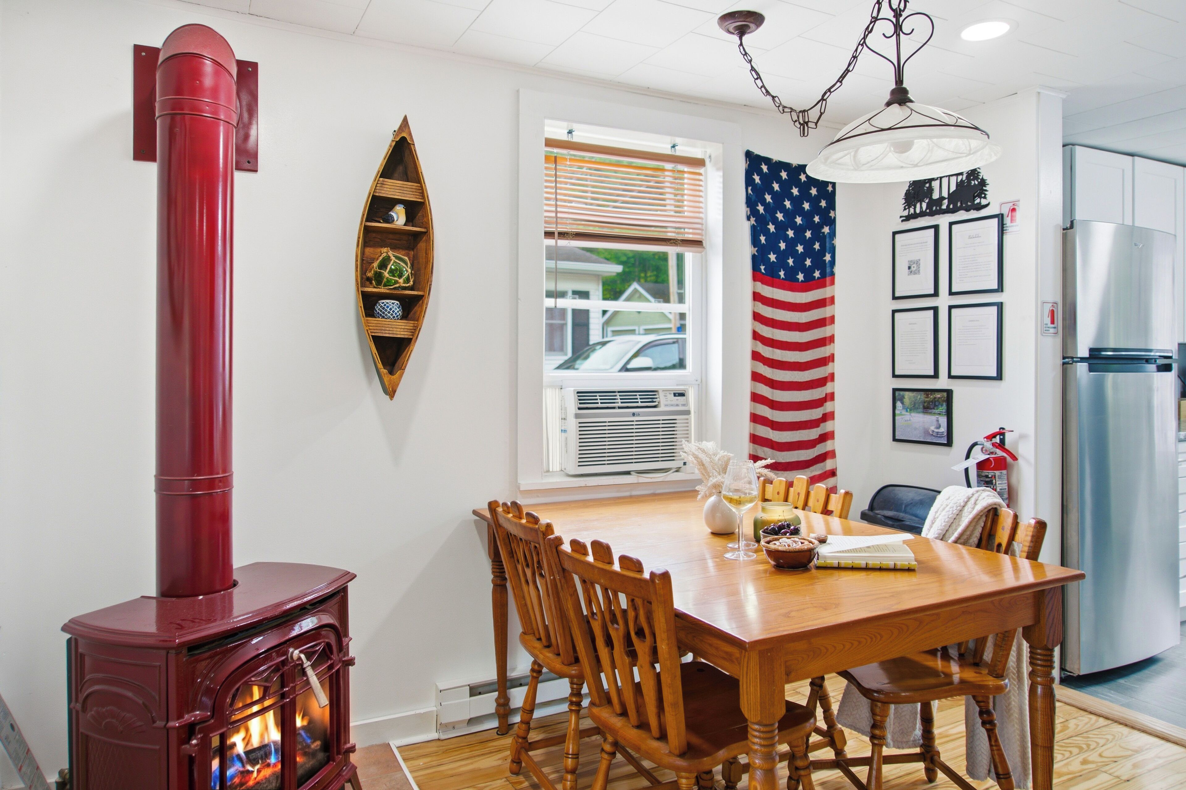 A dining area with a wooden table, chairs, a red wood stove, an American flag, and a refrigerator.