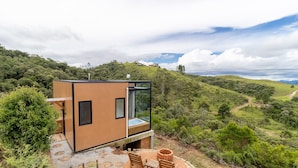 Una cabaña moderna y compacta con ventanas amplias, ubicada en una colina con vistas panorámicas del paisaje circundante.