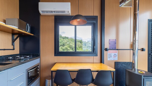 Cocina compacta con estufa a gas, horno y mesa de madera con sillas. Hay una ventana con vistas a zonas verdes y un cartel en la pared.
