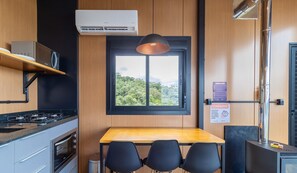 Cocina compacta con estufa a gas, horno y mesa de madera con sillas. Hay una ventana con vistas a zonas verdes y un cartel en la pared.